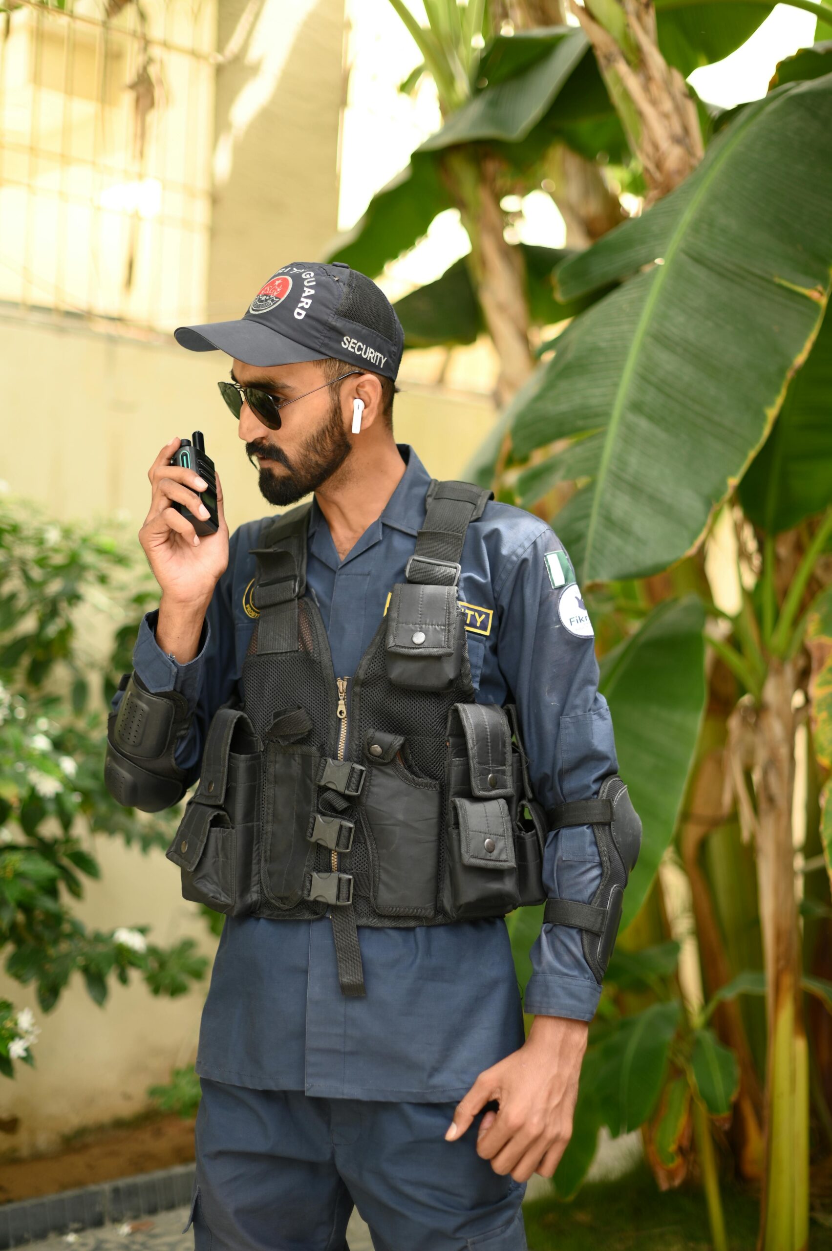 Security guard using walkie talkie in outdoor setting with greenery.